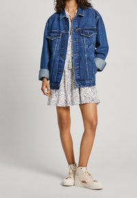 Jeanskjacka med lös passform, blå färg och framfickor, ihop med en blommig klänning ochbeige hög-top sneakers.