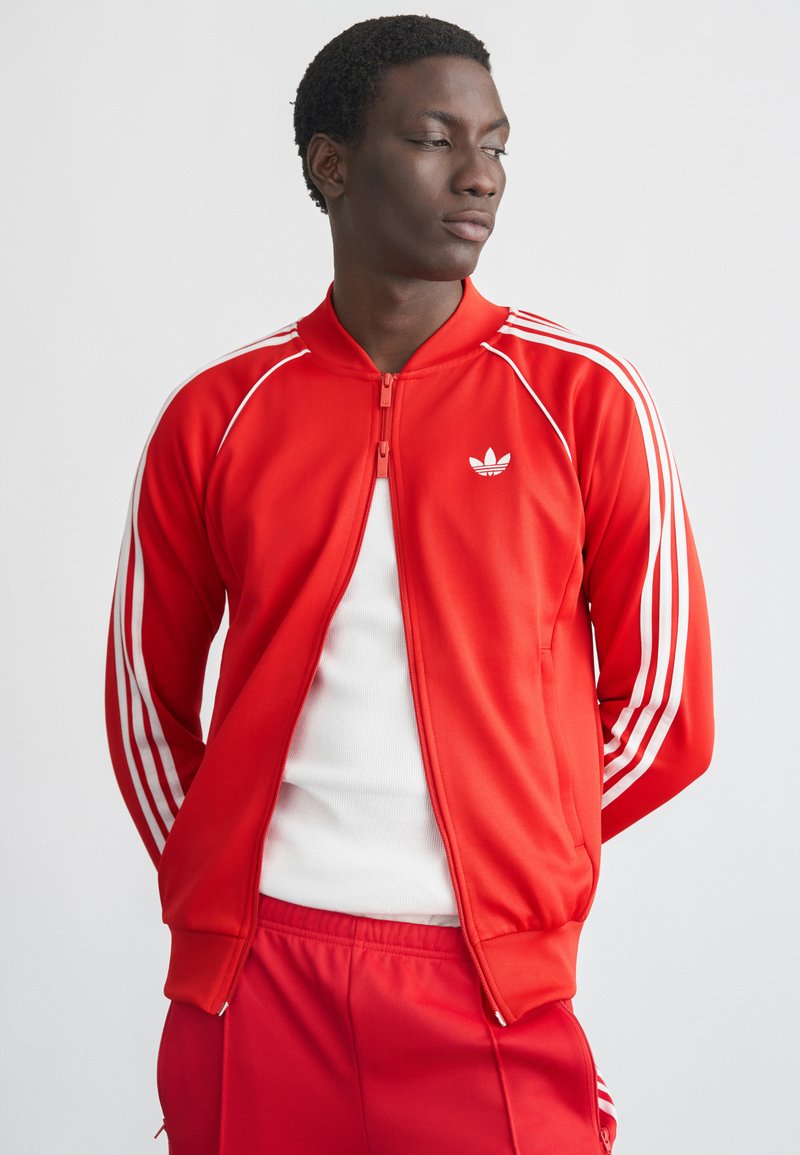 Casaco desportivo de poliéster vermelho com riscas brancas ao longo das mangas e fecho de correr frontal, apresentando uma gola redonda e um logótipo bordado no peito.