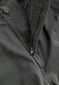Dunkelgraue Hose mit Reißverschluss und Knopfverschluss, sichtbaren Steppnähten und einer glatten, robusten Stoffstruktur.