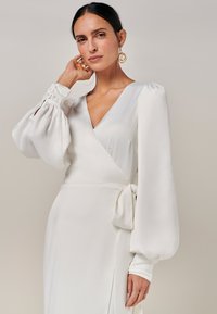 Weißes Wickelkleid mit langen Puffärmeln und gebundenem Taillenband. Glatte, seidige Textur mit tiefem V-Ausschnitt und seitlichem Schlitz. Goldene Creolen als Akzente.