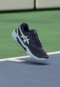 Marineblå treningssko med hvite detaljer, nettstoff på overdelen og gummisåle, vist svevende over en tennisbanes overflate.