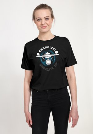 Femme portant un t-shirt noir avec un motif de vaisseau spatial et le texte « Energize ! Beam me up » sur un fond clair uni.
