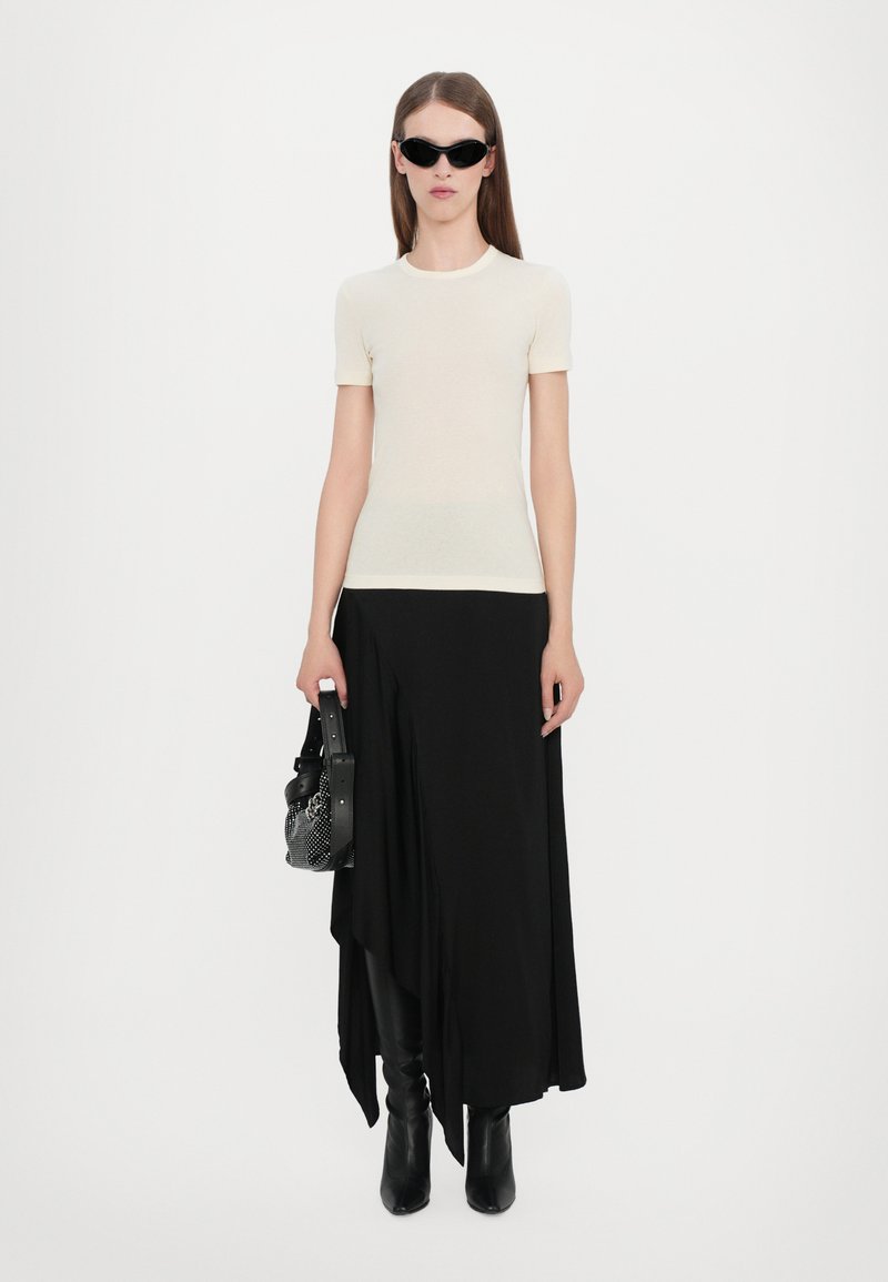 Top ajustado color crema claro, falda negra fluida con dobladillo asimétrico y botas negras hasta la rodilla. Sosteniendo un pequeño bolso negro y plateado.