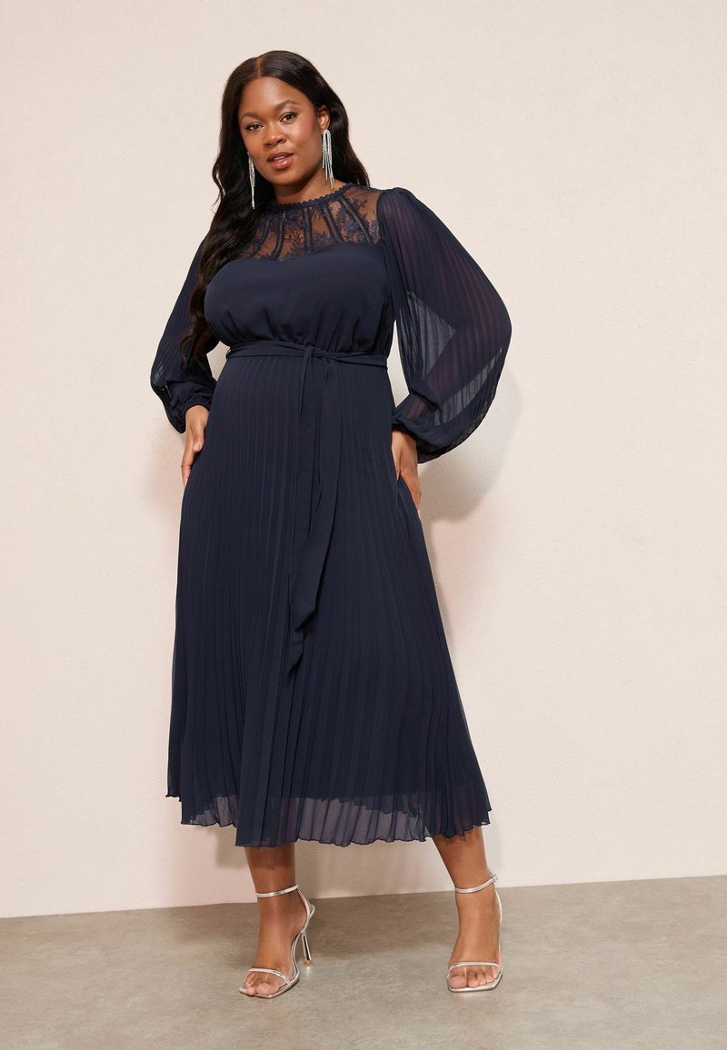 Tumes pleediga kleit, millel on läbipaistev pitsist ülemine osa, kõrge kaelus ja pikad, õhulised varrukad; vöökoha peal on kinni seotud sobiva vööga ja seda täiustavad rihmadega kingad.