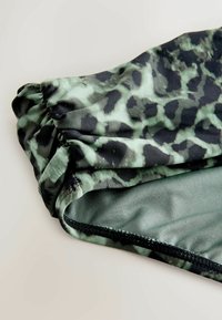 Vihreä ja musta leopardikuvioinen kangas, jolla on sileä pinta, kokoontaitetut reunat ja musta tikattu helma.