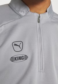 Vaaleanharmaa urheilutakki, jossa on puolivetoketju, mustalla Puma-logolla ja "KING"-tekstillä alla, tekstuuri, jossa on hienovarainen kuvio.