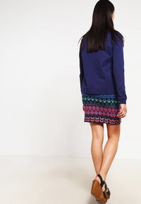 Donna di spalle che indossa un maglione blu navy, una gonna di maglia a fantasia multicolore e sandali con zeppa neri con suola in legno.