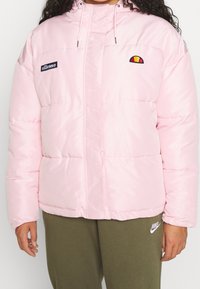 Osoba v světle růžové prošívané bundě s logy ellesse na hrudi a olivově zelených teplácích Nike.