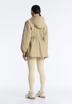 Femeie văzută de spate purtând o jachetă matlasată bej cu glugă, colanți crem, șosete albe și adidași bej pe fundal alb.