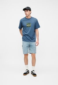 Νεαρός άνδρας με μπλε ναυτικό t-shirt με σχέδιο, ανοιχτόχρωμα σορτς τζιν, μαύρα αθλητικά παπούτσια, λευκές κάλτσες και μαύρο καπέλο μπέιζμπολ με ρίγες, στέκεται σε λευκό φόντο.