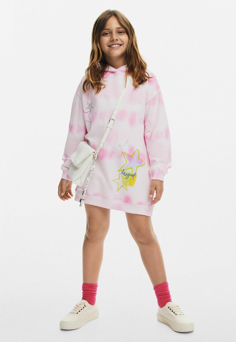 Robe sweat en tie-dye rose avec des graphiques d'étoiles en jaune et argent. Associée à des baskets blanches et des chaussettes montantes roses, avec un sac bandoulière blanc.