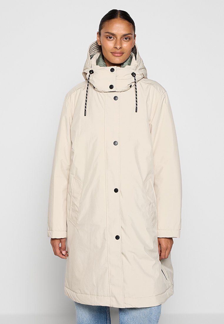 Femme portant un long manteau d'hiver beige à capuche, avec des boutons noirs et des poches latérales, associé à un jean bleu clair.