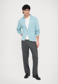 Ανοιχτό μπλε ριμπ πλεκτό πουλόβερ με φερμουάρ πάνω από λευκό T-shirt, συνδυασμένο με γκρι πλισέ παντελόνι και μαύρα παπούτσια. Το μοντέλο στέκεται μπροστά σε λευκό φόντο.