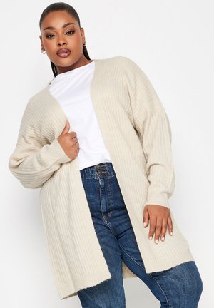 Licht beige geribde cardigan met een losse pasvorm, open voorkant en lange mouwen, gecombineerd met een wit T-shirt en donkerblauwe jeans.
