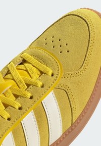 Gul mockasneaker med vita läderstripes, utrustad med gula skosnören och perforerad detaljering på tån, placerad på en brun gummisula.