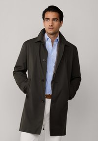 Tumma oliivinvihreä trench-coat, jossa on nappikiinnitys, kaulus ja sivutaskut, kerroksittain vaaleansinisenvivahteisen raidallisen paidan ja vaaleiden housujen päällä.