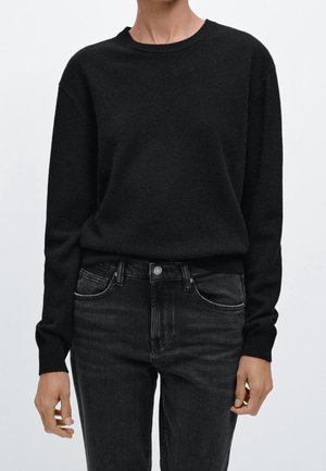 Persona che indossa un maglione nero a maniche lunghe semplice infilato in un paio di jeans neri slavati a vita alta, in piedi contro uno sfondo bianco uniforme.