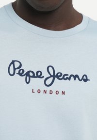 Gaiši zils īsām piedurknēm T-krekliņš ar apaļu kakla ārmalu, kurā ir tumši zila teksts "Pepe Jeans" un sarkans "LONDON" zem tā ikdienišķā fontā.