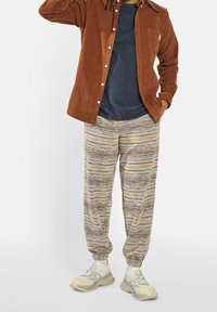 Camisa de pana marrón, camiseta azul marino, pantalones jogger beige con estampado, zapatillas grises. Detalles notables: rayas horizontales y puños elásticos en los pantalones.