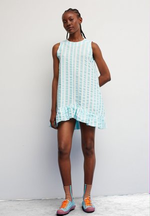 Young woman with braided hair wearing a sleeveless blue and white striped dress with ruffled hem, colorful socks, and sneakers standing against a white wall.