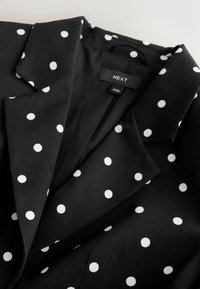 Blazer noir à pois blancs, avec un col à revers cranté et une étiquette de vêtement portant la mention "NEXT" à l'intérieur de l'encolure.