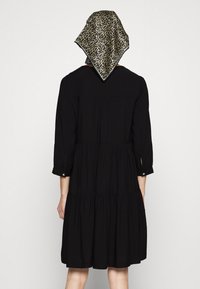 Schwarzes, schichtweise gestaltetes Kleid mit Dreiviertelärmeln, das mit einem Knopfdetail versehen ist. Ein Leopardenmuster-Schal ist am Hinterkopf gebunden.