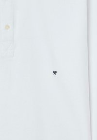 Polo blanca hecha de algodón, con una tapeta de tres botones, un pequeño logo negro bordado en el lado izquierdo del pecho y una tela texturizada.