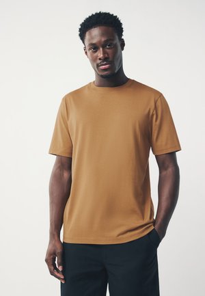 Mladý muž s krátkymi kučeravými vlasmi, oblečený v jednofarebné hnedé tričko s krátkym rukávom a tmavé nohavice, stojaci na svetlom pozadí.