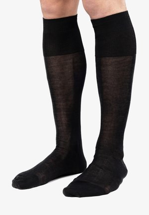 Benen met zwarte doorschijnende kniehoge sokken met een stevige geribde boord tegen een witte achtergrond.