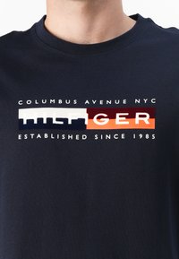 Mørkeblå bomulls t-skjorte med fet hvit og farget tekstgrafikk som leser "COLUMBUS AVENUE NYC HILFIGER ETABLERT SIDEN 1985."