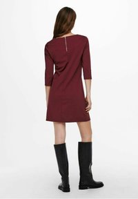 Robe bordeaux à manches trois-quarts, avec une fermeture éclair au dos et une coupe droite, associée à des bottes noires montantes jusqu'aux genoux.
