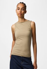 Jeune femme aux cheveux tressés portant un haut beige sans manches et un jean bleu foncé, debout sur un fond blanc uni.