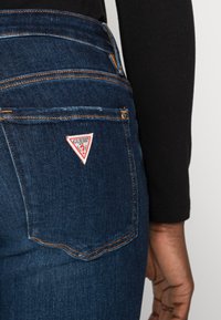 Un bolsillo de jeans de mezclilla azul oscuro con costuras naranjas y un parche blanco con el logo de Guess en la parte trasera, junto a la mano de una persona y una manga negra.
