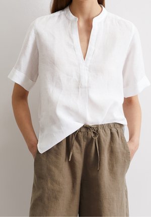 Persoon die een witte blouse met korte mouwen en V-hals draagt en bruine linnen broek met trekkoord, met handen in de zakken tegen een effen achtergrond.