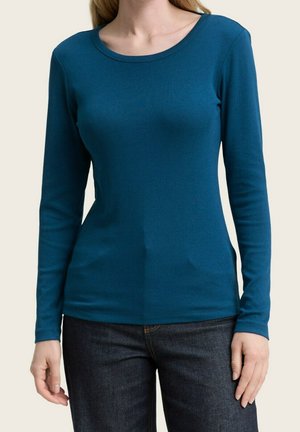 Femme portant un haut ajusté à manches longues couleur sarcelle et un jean bleu foncé, debout devant un fond clair uni.