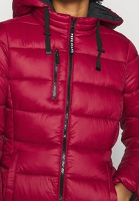 Chaqueta acolchada roja tipo puffer con cremallera negra, cremallera en el bolsillo del pecho, capucha con cordones negros y texto "PEPE JEANS" en el tirador de la cremallera principal.