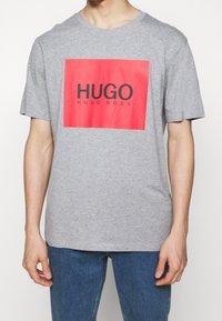 Camiseta de manga corta gris hecha de algodón, con un gran gráfico rectangular rojo que presenta el texto "HUGO" y "HUGO BOSS" en negro.
