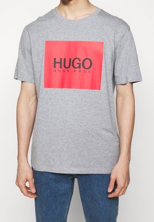 T-shirt gris à manches courtes en coton, arborant un grand graphique rectangulaire rouge avec le texte "HUGO" et "HUGO BOSS" en noir.