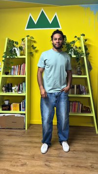 Άνδρας με ανοιχτό μπλε μπλουζάκι και τζιν στέκεται μπροστά από έναν κίτρινο τοίχο με πράσινο τρίγωνο, ανάμεσα σε δύο πράσινα ράφια με φυτά και βιβλία.