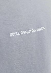 Pelēks audums ar balto izšūto tekstu "ROYAL DENIM DIVISION". Faktūra ir gluda ar vieglu spīdumu.