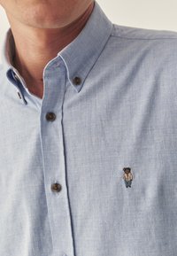 Cămașă cu nasturi de culoare albastru deschis, confecționată dintr-un material moale. Dispune de nasturi maro și un logo brodat cu un urs pe partea stângă a pieptului.