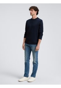 Camicia a maniche lunghe blu navy in tessuto morbido, abbinata a jeans azzurri e sneakers bianche. Modella in una posa neutra contro uno sfondo semplice.