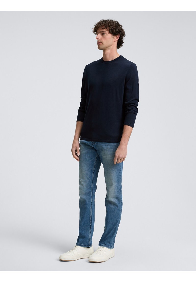 Camicia a maniche lunghe blu navy in tessuto morbido, abbinata a jeans azzurri e sneakers bianche. Modella in una posa neutra contro uno sfondo semplice.