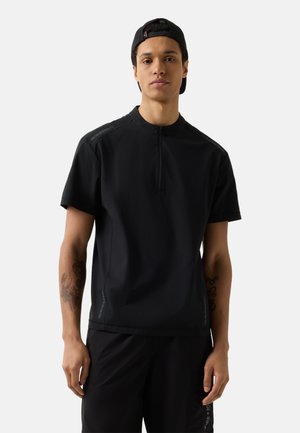 Jeune homme portant une chemise zippée noire à manches courtes, un short noir et une casquette noire portée à l'envers, debout devant un fond uni.