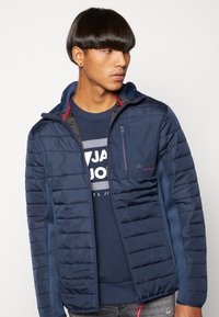 Marineblauwe pufferjack met horizontaal gequilt textuur, mesh voering, hoge kraag en een ritszak op de borst. Gelaagd over een donkerblauw sweatshirt.
