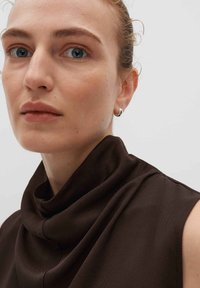 Top marrone con collo a cowl realizzato in tessuto liscio, caratterizzato da una texture drappeggiata e dettagli di cucitura minimi. Sono visibili piccoli orecchini a cerchio in argento.