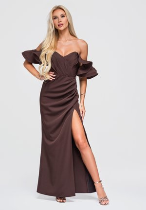 Femme aux longs cheveux blonds portant une robe bustier marron foncé avec des manches à volants et une fente haute, posant avec une main sur la hanche.