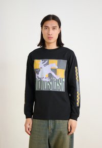 Schwarzes Langarmshirt mit einem grafischen Druck in Lila, Gelb und Grau. Lockere Passform mit karierter Musterung an den Ärmeln. Aus weichem Baumwollstoff.