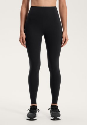 Leggings neri a vita alta realizzati in tessuto elastico, con una texture liscia e un design senza cuciture. Indossati con scarpe da ginnastica nere.
