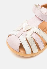 Svijetlopinkane suede sandale s bijelim i srebrnim detaljima, otvorenim prstima, detaljem u obliku srca i podesivom trakom. Ravna potplata.
